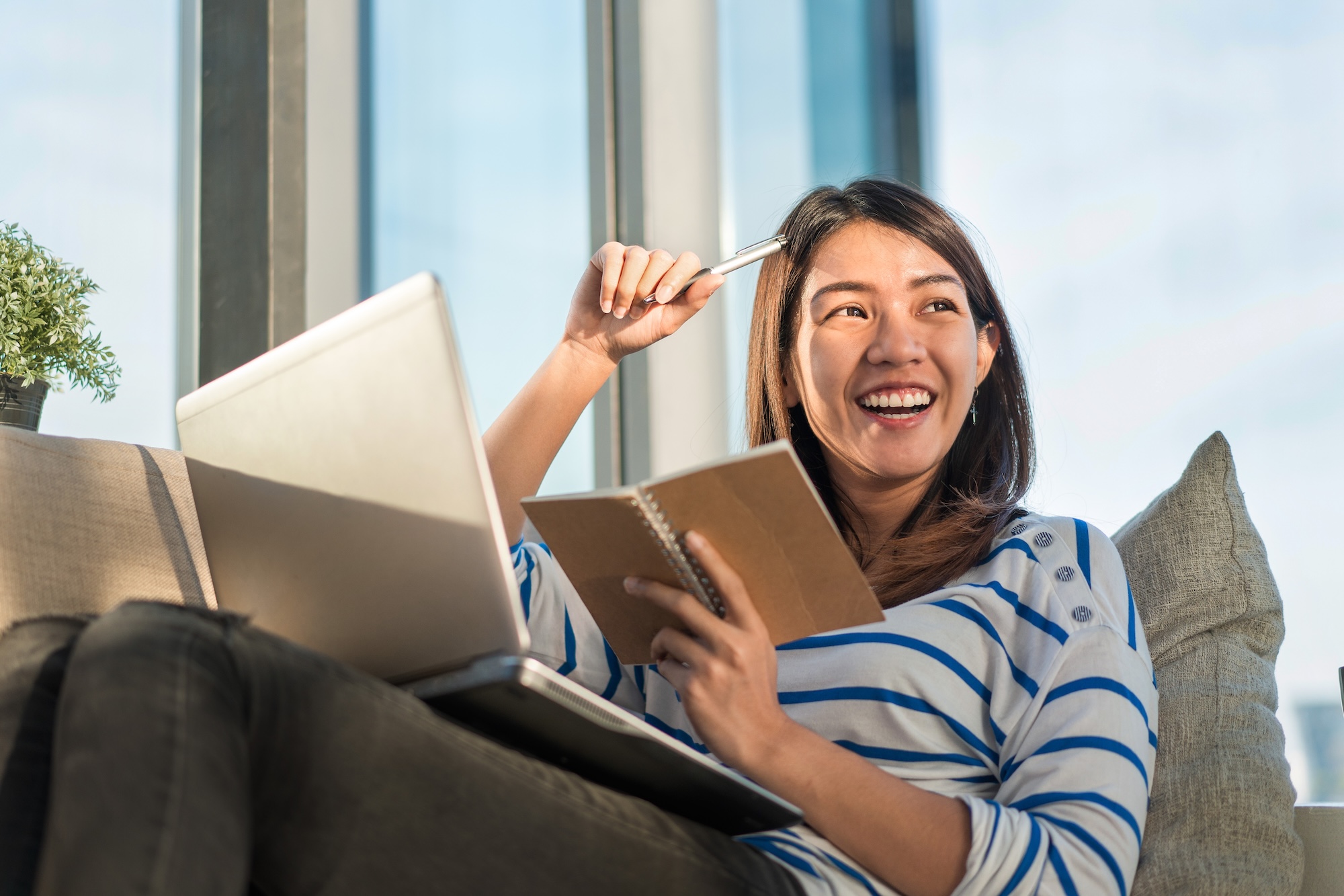 Happy smile young asian woman working with laptop and hold pen with notebook and sitting on sofa at home office, Selective focus
